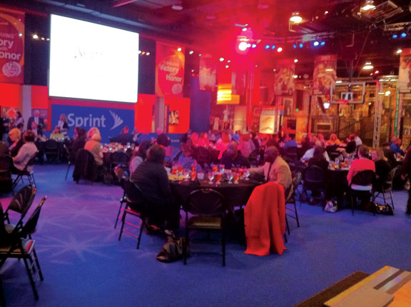 Conference event with attendees seated at round tables, colorful lighting, and a large presentation screen.