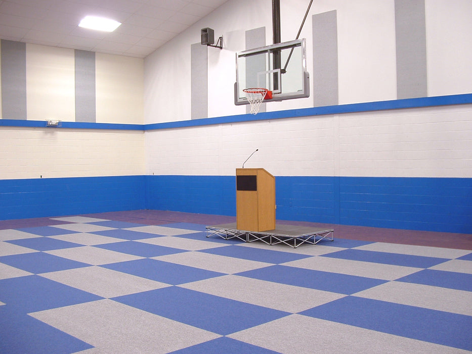 Spacious gym with blue and gray checkered flooring, podium for presentations, basketball hoop in background.