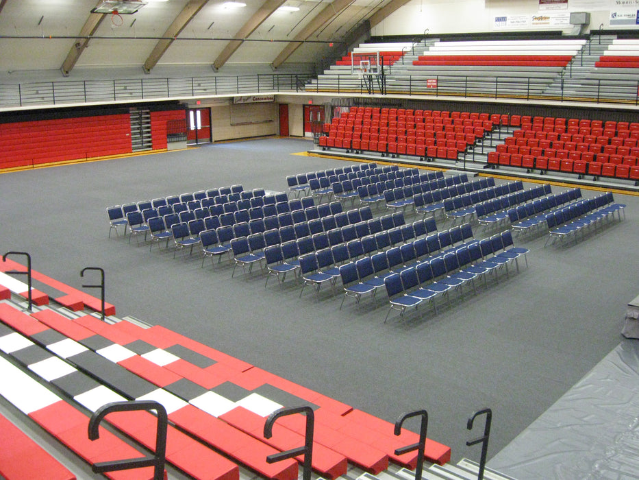 Rows of blue folding chairs in a spacious gymnasium with red and black bleachers.